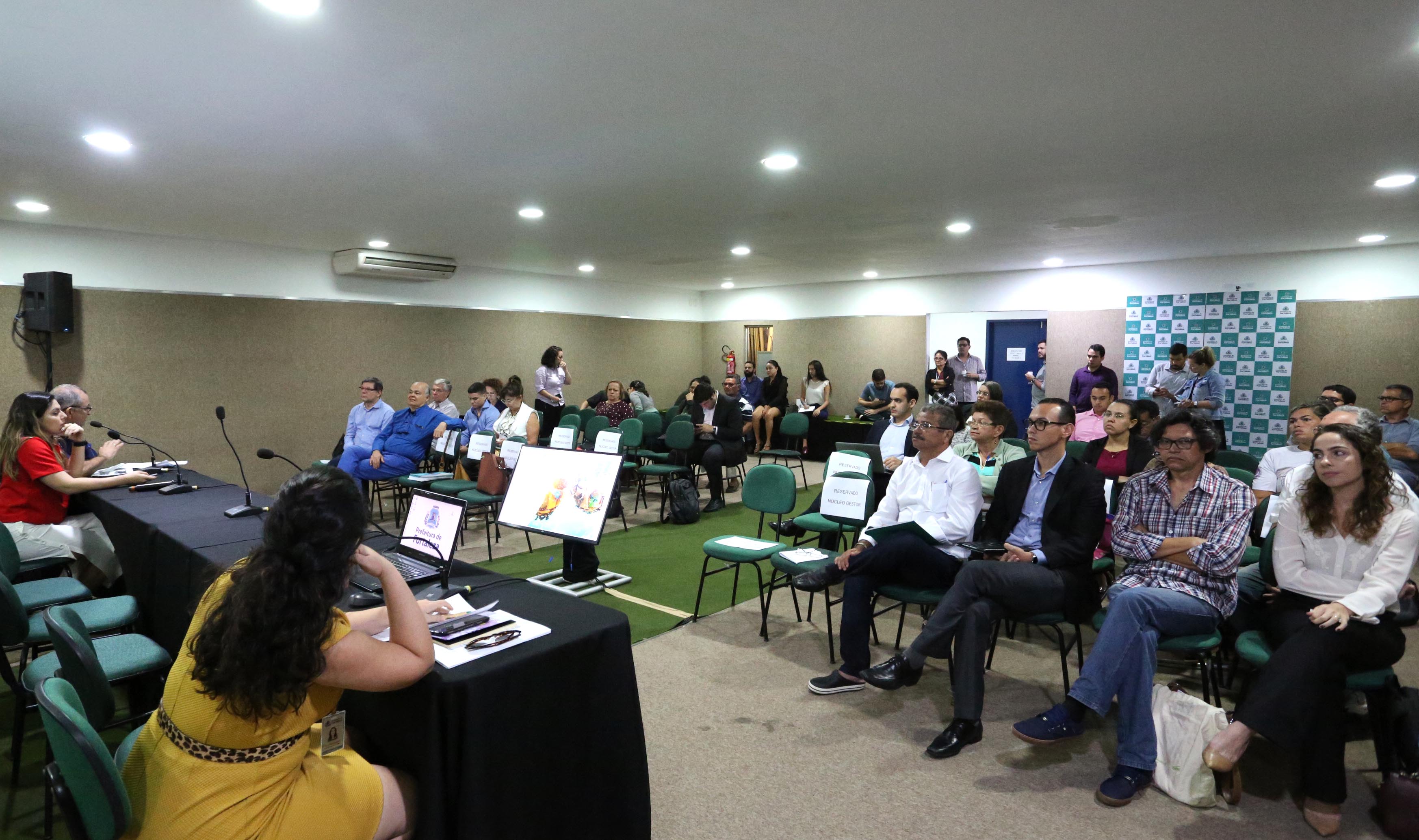 pessoas sentadas em plateia em frente à mesa com pessoas falando